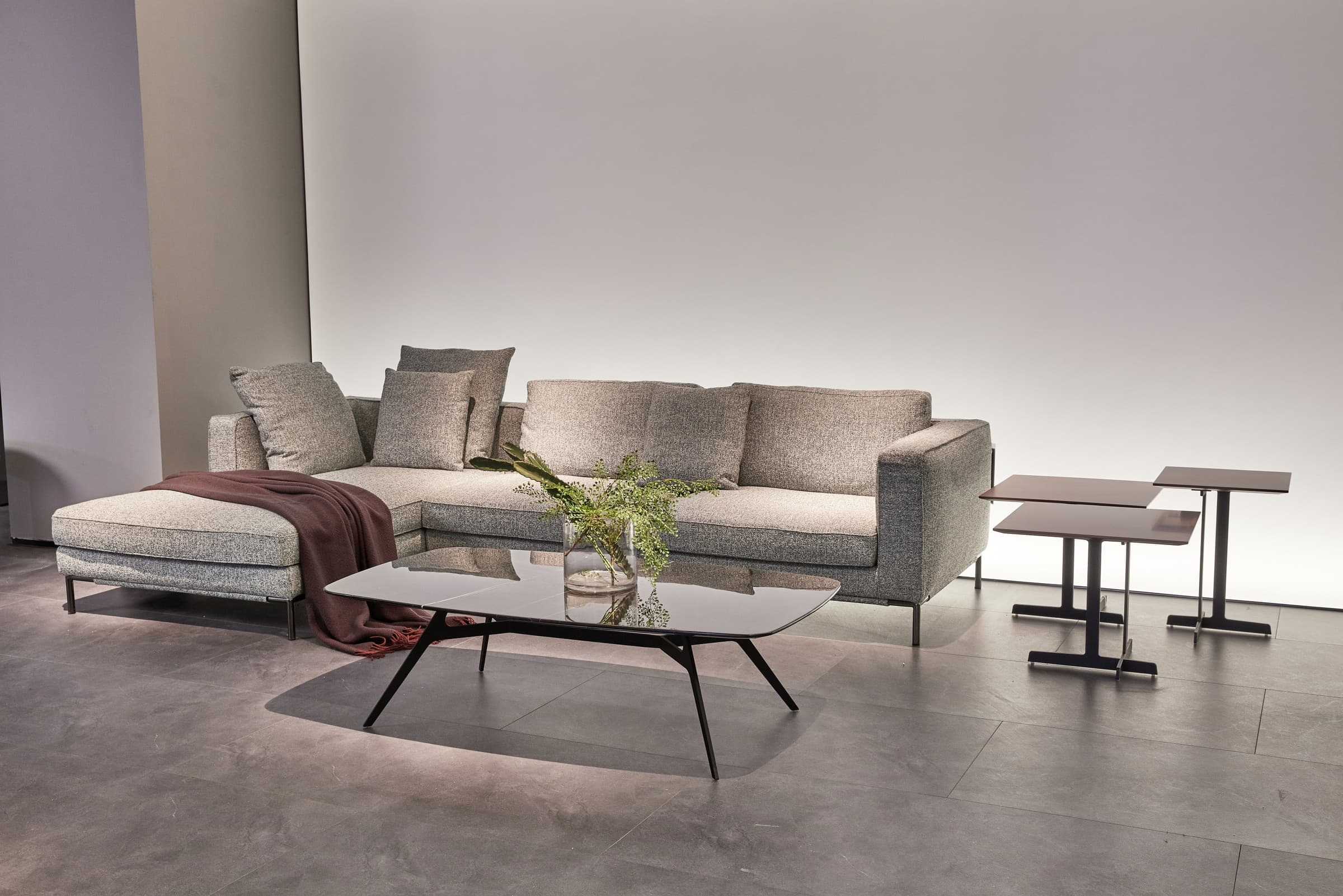 Rein chaise sofa in oat-grey colorway with stacked back cushions and a brown throw beside an angular glass coffee table
